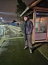 Mann i mørke klær står ved en opplyst bussholdeplass en våt kveld.